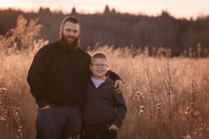 A bearded dad and his son stand together with arms around each other.