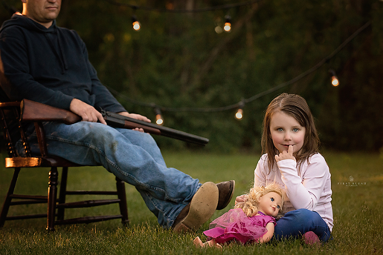 Little girl smiles and sits on the grass by her watchful father near Red Deer Alberta.