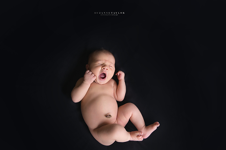 Baby boy yawns while naked on a black backdrop