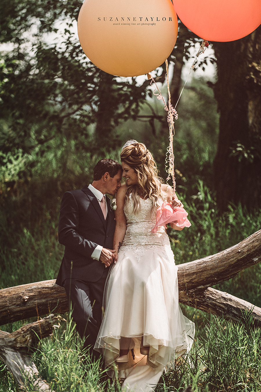 A newly married couple sits on a fallen tree together while the groom kisses her shoulder holding balloons during their Calgary Wedding.