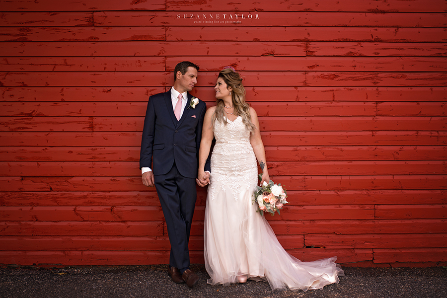 A newly married couple stands holding hands against a red wooden wall during their Calgary Wedding.